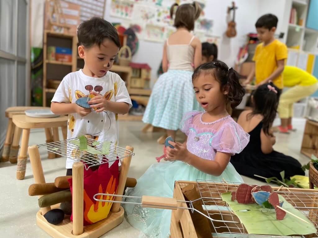 เด็กสองคนกำลังเล่นของเล่นไม้สีสันสดใสอย่างมีความสุข ภายในมุมเล่นที่สว่างไสว เต็มไปด้วยเบาะนุ่มและการตกแต่งที่สดใสร่าเริง