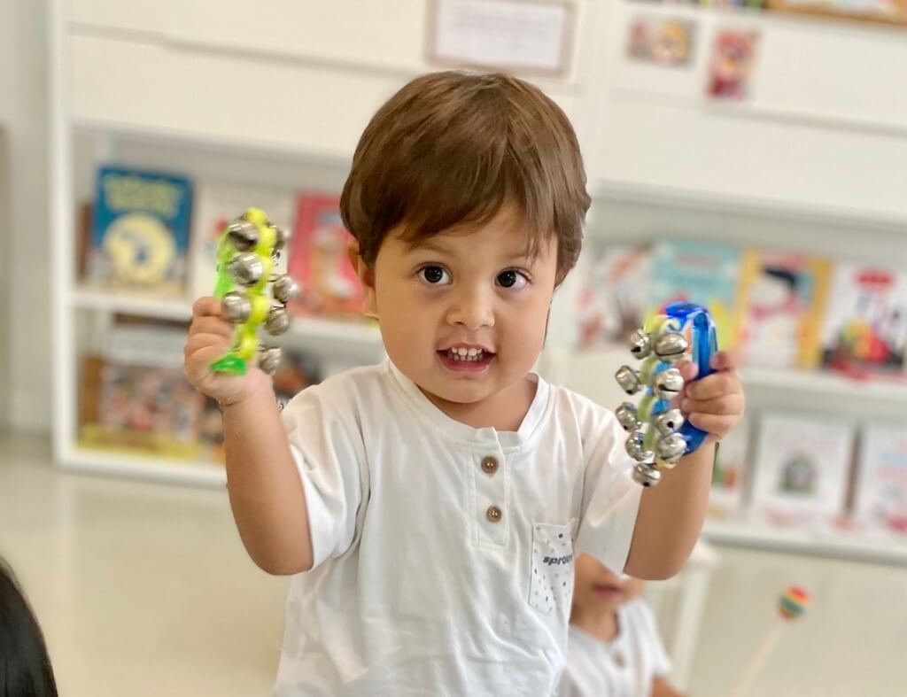 เด็กชายตัวน้อยยกของเล่นสีสันสดใสสองชิ้นขึ้นอย่างภาคภูมิใจ ภายในห้องเรียนที่สว่างสดใส