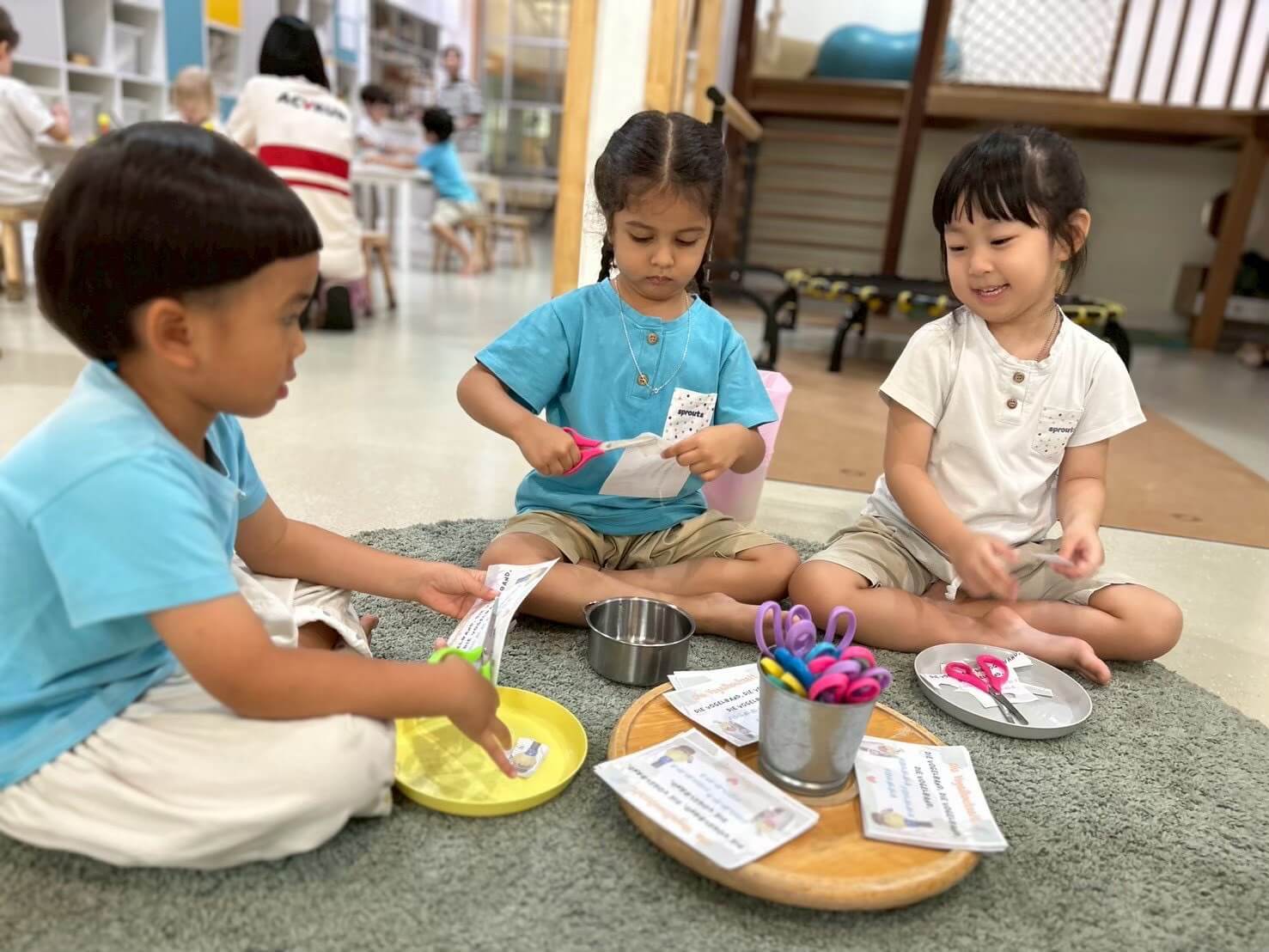 เด็กสามคนนั่งอยู่บนพื้น กำลังเล่นอย่างสนุกสนานท่ามกลางของเล่นหลากสีที่กระจายอยู่รอบตัว